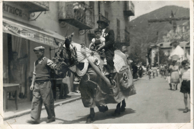 Photo d'archives représentant un village de l'arrière pays maralpin. Un cheval décoré est guidé par un villageois tandis qu'il porte sur son dos un enfant et un adulte